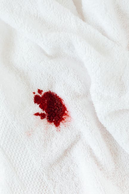 Close-up of a white fabric surface, likely a bedsheet or towel, with a small, irregularly shaped fresh bloodstain positioned towards the lower left. The fabric appears clean with a soft, textured appe